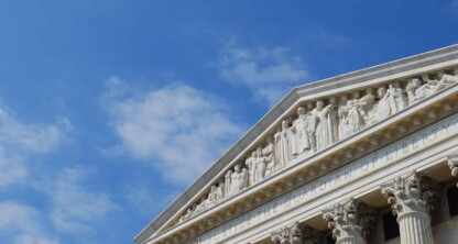 Partial view of a courthouse facade, reminiscent of prestigious law offices, with grand columns and sculpted figures under a blue sky with scattered clouds.