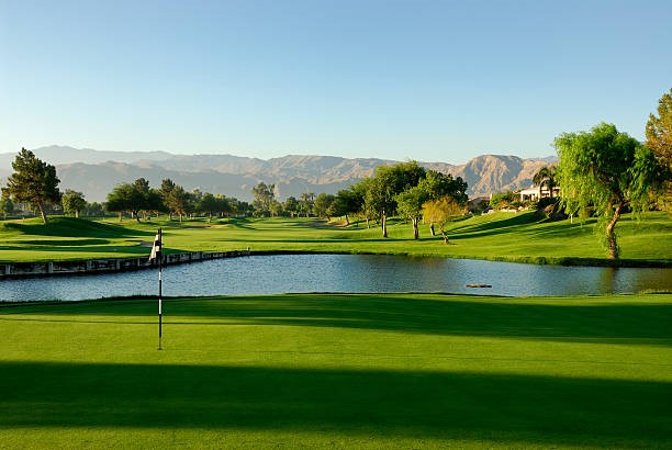 A golf course with a putting green, water hazard, trees, and mountains in the background under a clear blue sky—an ideal retreat for lawyers in Chicago after a busy day at the corporate law office.
