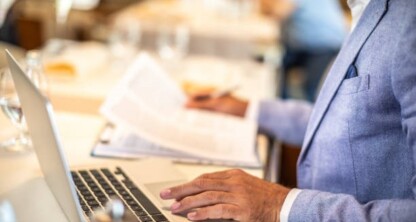A person in a light blue blazer works at a table with a laptop and documents, holding papers in one hand and typing with the other, capturing the busy atmosphere of a corporate law office or lawyers in Chicago.