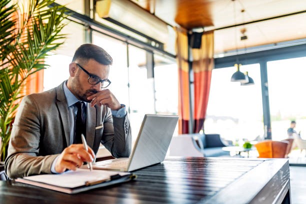 Un hombre vestido con traje está sentado a una mesa en una cafetería moderna, trabajando con un ordenador portátil y tomando notas en un cuaderno; quizá esté preparando material de apoyo para un litigio destinado a destacados abogados de Chicago.