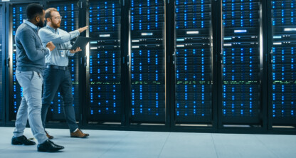 Dos hombres están de pie en una sala de servidores, uno de ellos sostiene un ordenador portátil y gesticula mientras hablan sobre el equipo del centro de datos, destacando la tecnología que utilizan los abogados de Chicago para el apoyo en litigios.