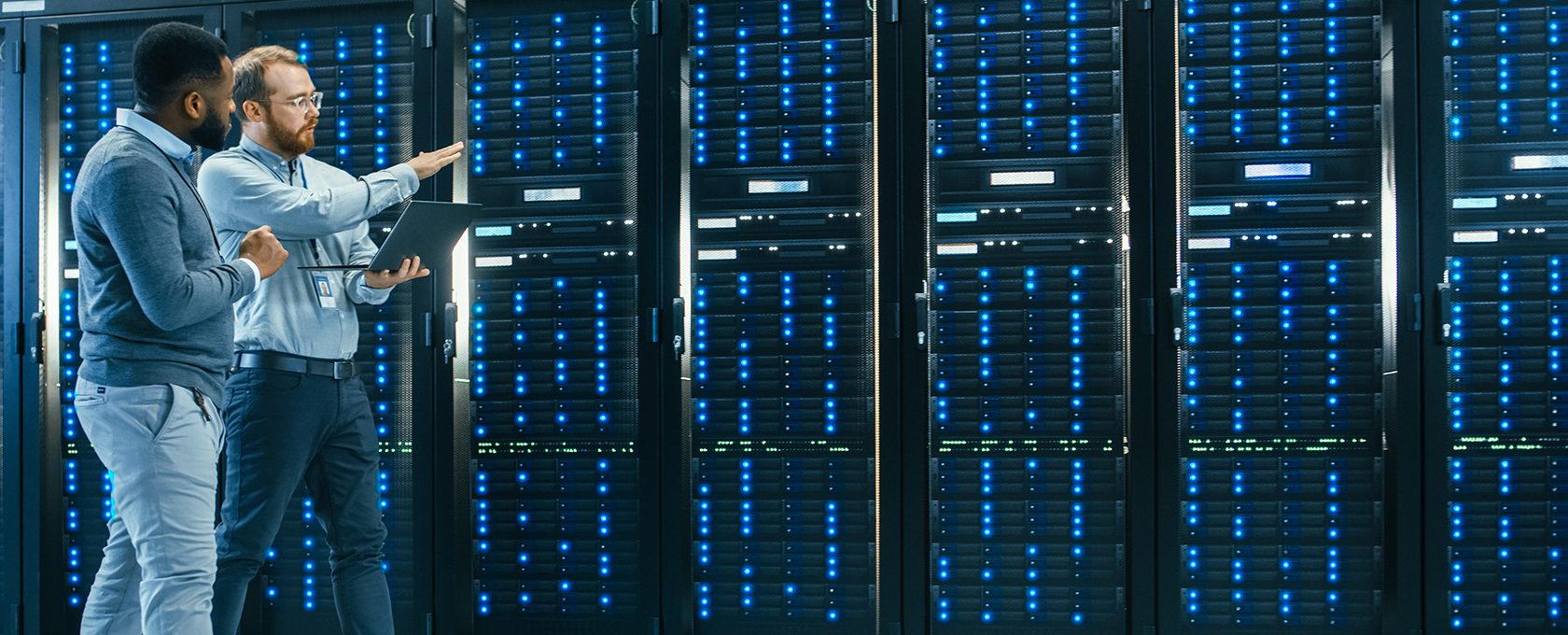 Two men stand in a server room with rows of server racks, one holding a laptop and pointing at the servers while discussing solutions for intellectual property law management.