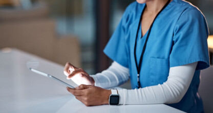 A person in blue medical scrubs uses a tablet at a desk, smiling, with a smartwatch on their wrist, as if consulting litigation support at a corporate law office.