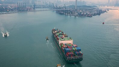 A large cargo ship carrying shipping containers enters a busy port, escorted by smaller boats, with a city skyline and mountains in the background—much like lawyers in Chicago navigating complex intellectual property law cases.
