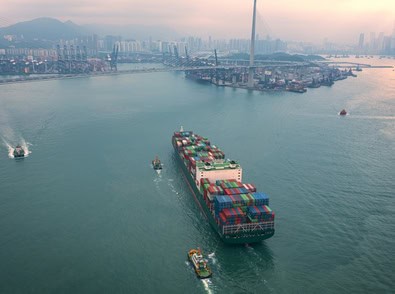 A large cargo ship carrying shipping containers enters a busy port, escorted by smaller boats, with a city skyline and mountains in the background—much like lawyers in Chicago navigating complex intellectual property law cases.