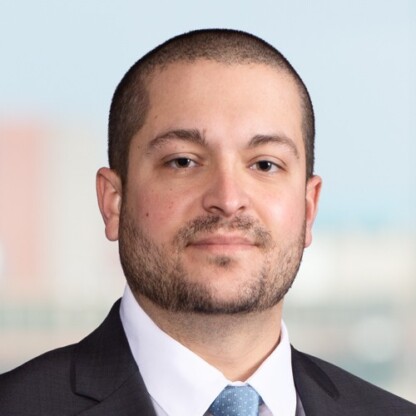 A man with short hair and a trimmed beard, dressed in a dark suit, white shirt, and light blue tie, poses in front of a blurred background, representing chicago lawyers specializing in intellectual property law.