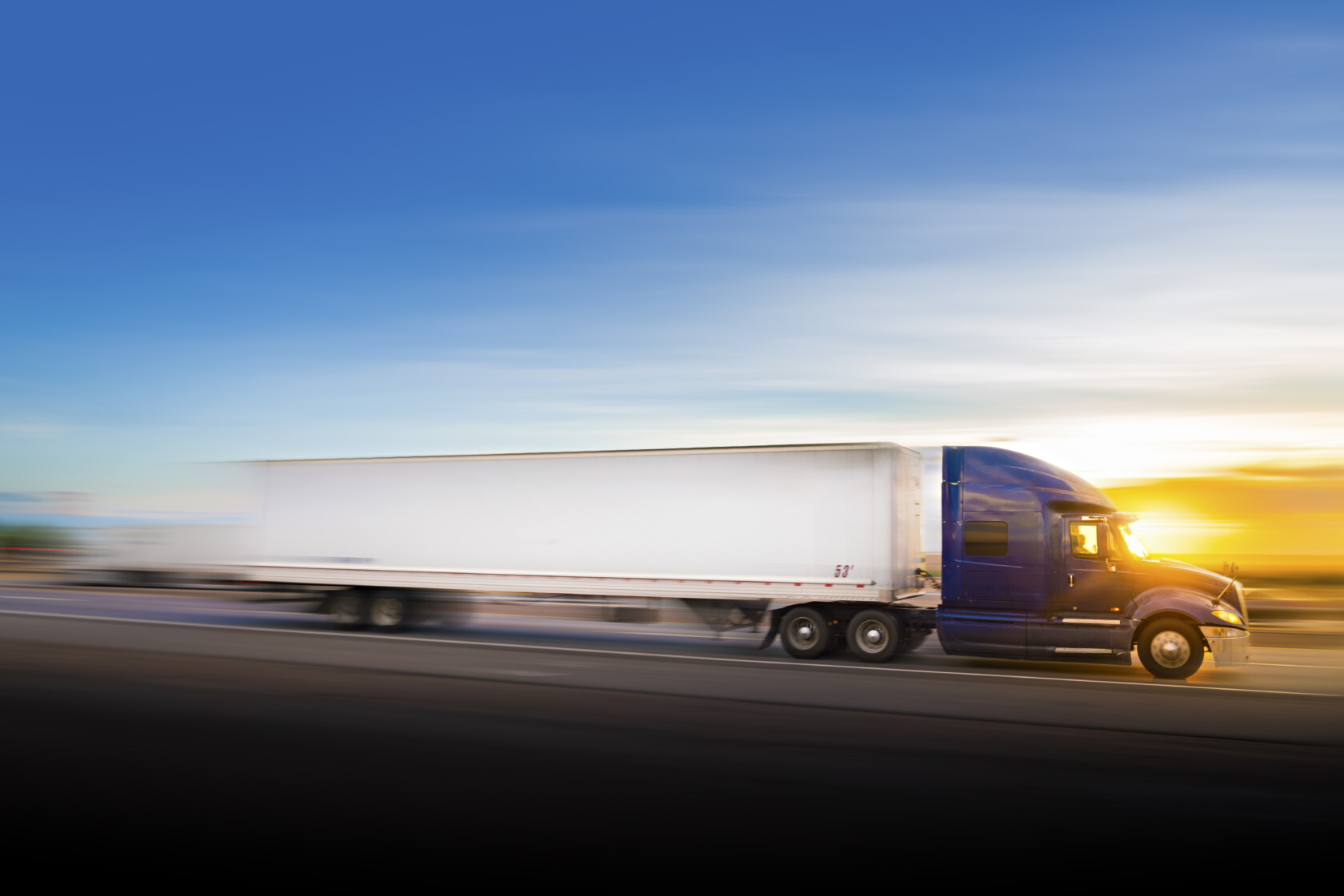 A blue semi-truck with a white trailer drives quickly on a highway at sunset, with a blurred background indicating motion.