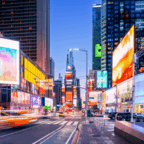 Times Square in New York City at dusk, with bright billboards, tall buildings, and blurred motion from traffic on the street.