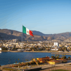Large Mexican flag on a flagpole near the waterfront with a city, mountains, and clear sky in the background.