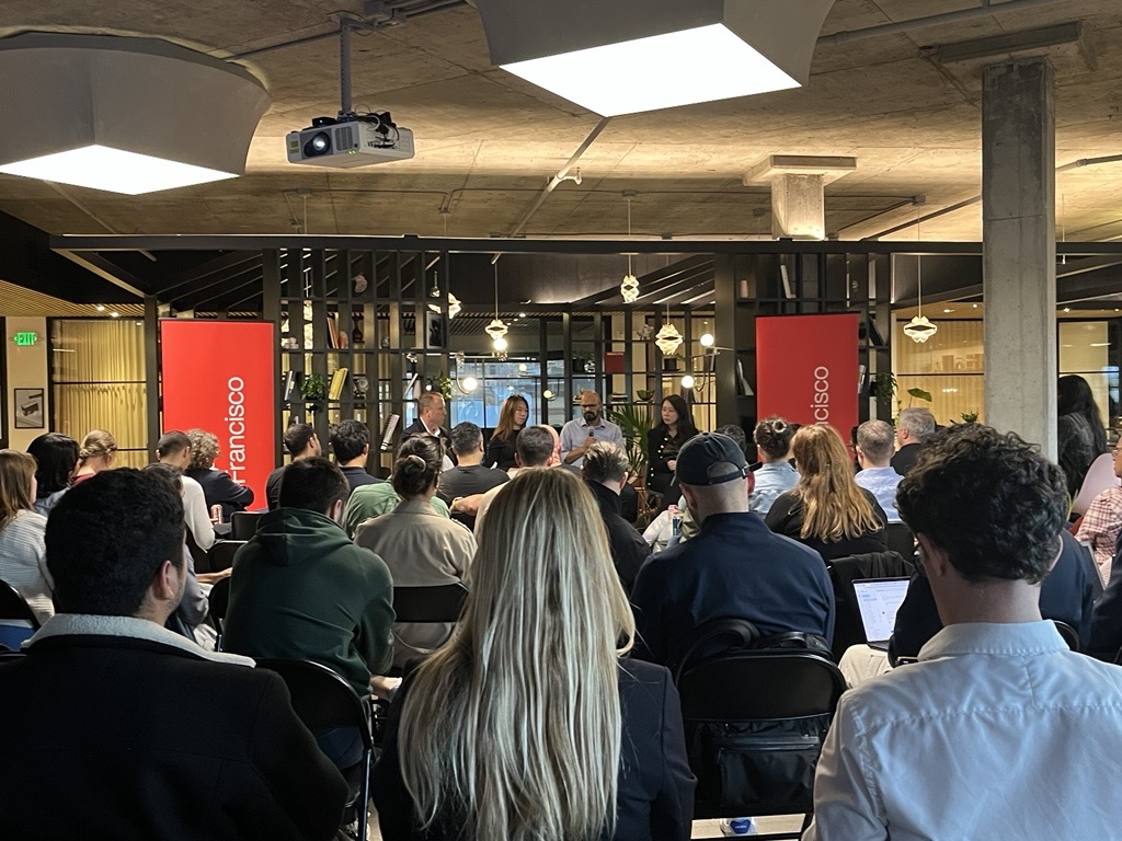 A group of people sits facing a panel of speakers at an indoor event, with two red "San Francisco" banners visible in the background.
