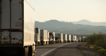 A long line of semi-trailer trucks is stopped along a highway with mountains visible in the background under a clear sky.