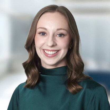 A woman with light skin and brown shoulder-length hair, wearing a dark green top, smiling at the camera against a blurred indoor background.