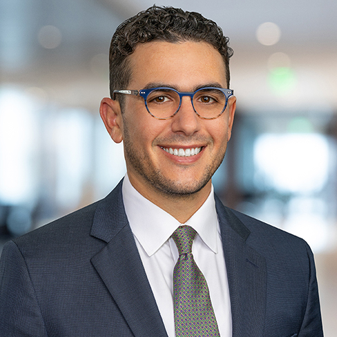Man wearing glasses, a dark suit, white shirt, and patterned tie, smiling in a brightly lit office setting.