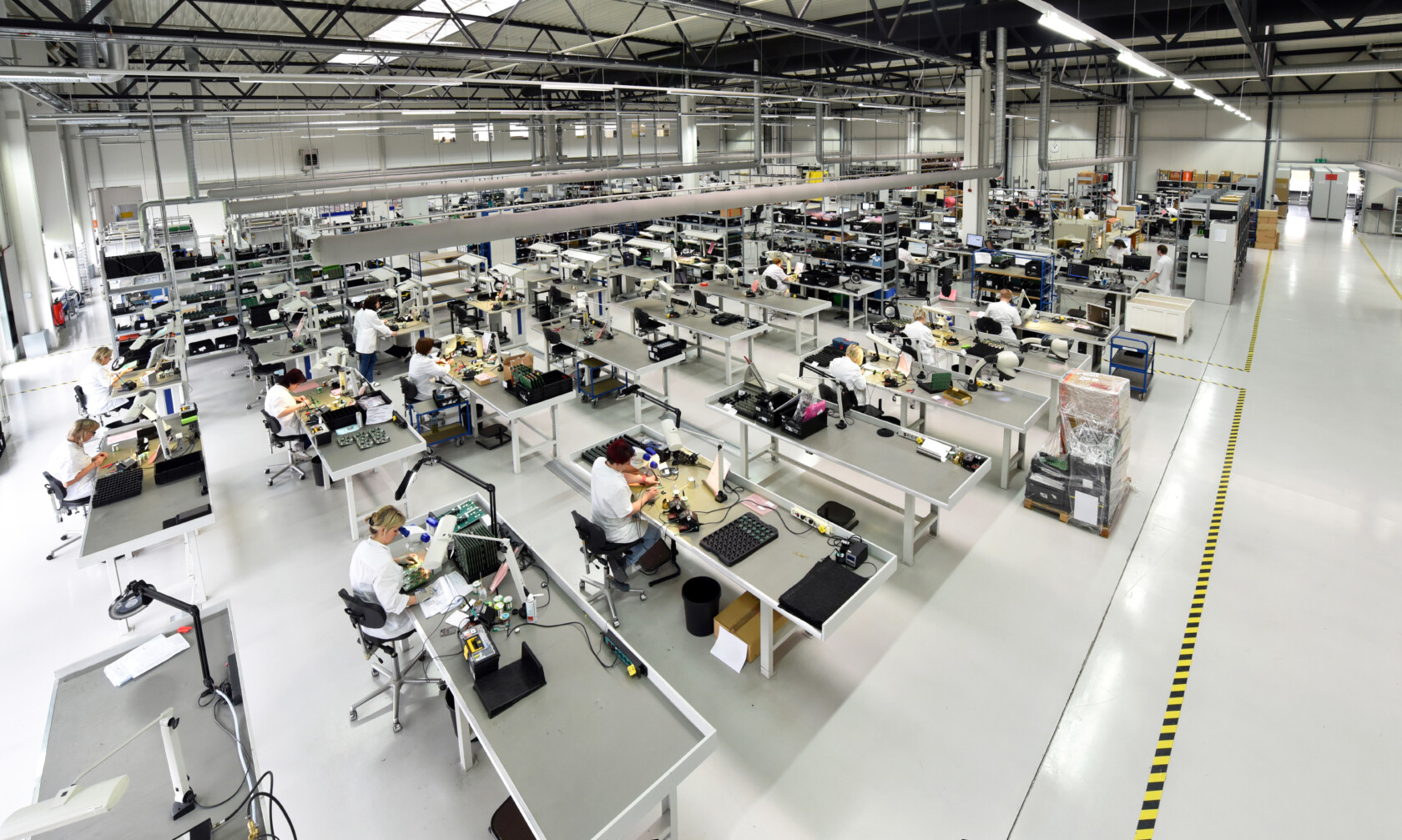 Large, brightly lit factory floor with multiple workstations where people assemble electronic components at long tables, surrounded by industrial equipment and supplies.