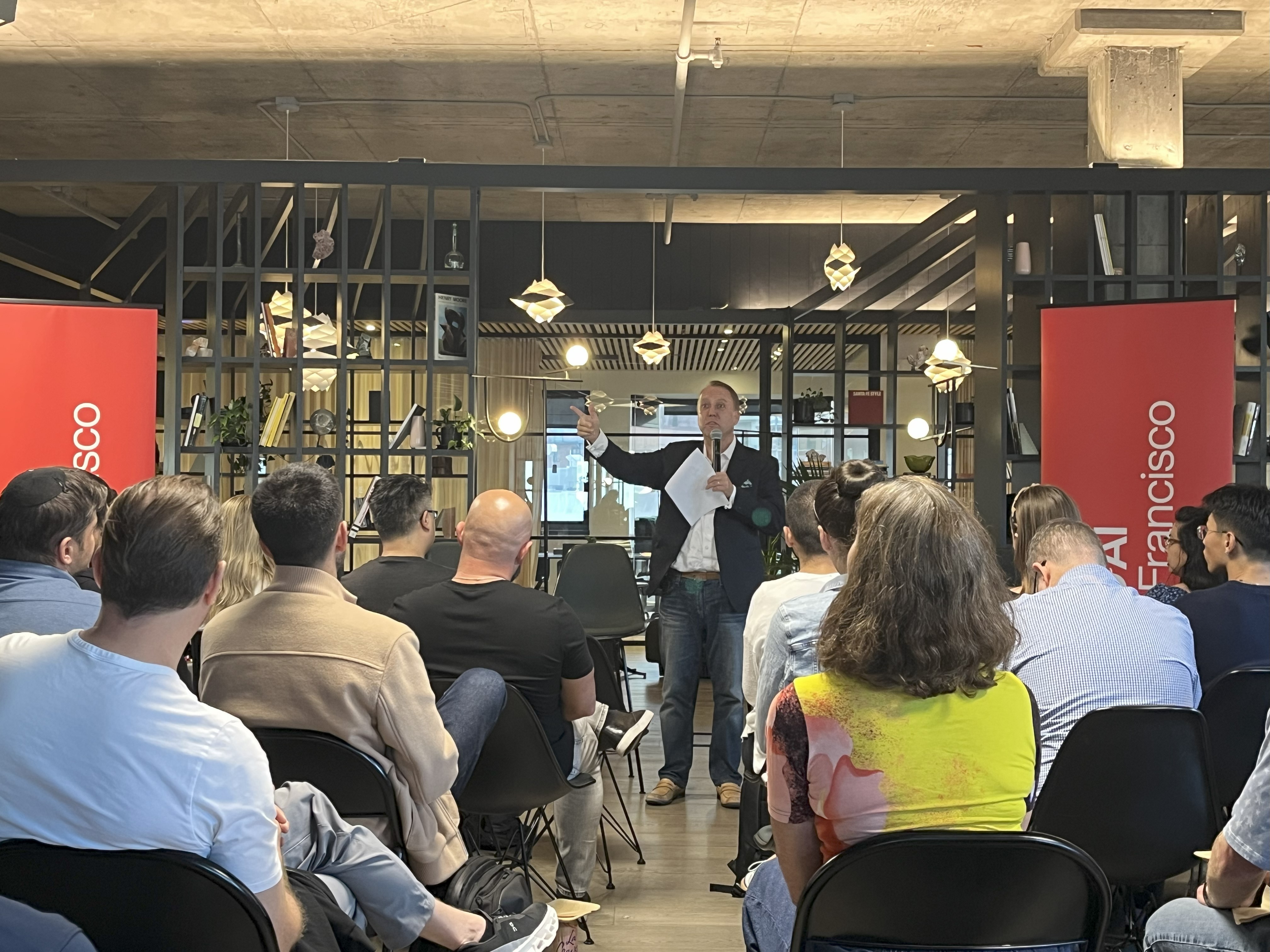 A man stands and speaks to an audience seated in rows at an indoor event, holding papers; two red banners read "San Francisco" on each side of the room.