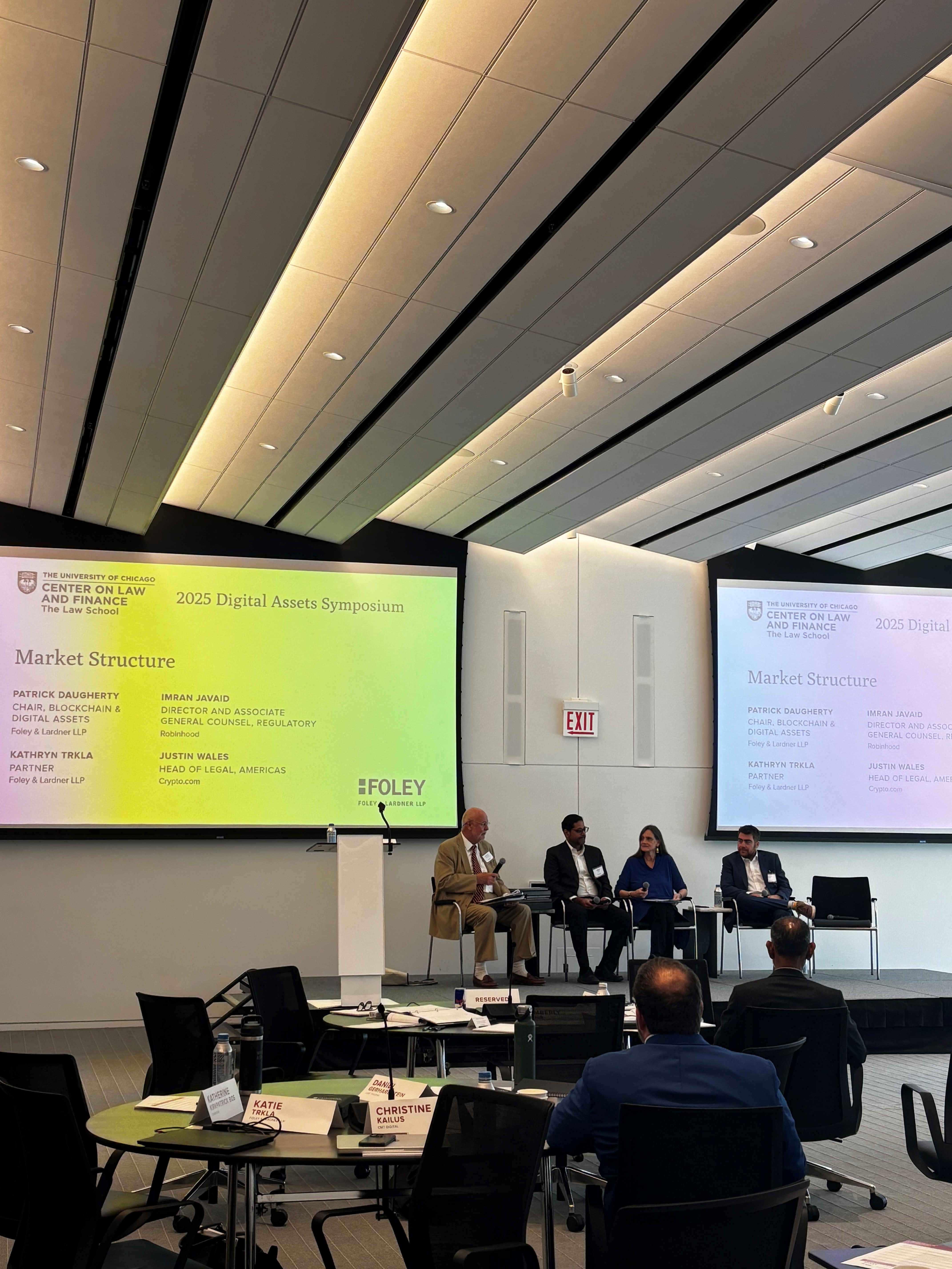 Four panelists sit on stage discussing "Market Structure" at the 2025 Digital Assets Symposium, with presentations displayed on large screens behind them.