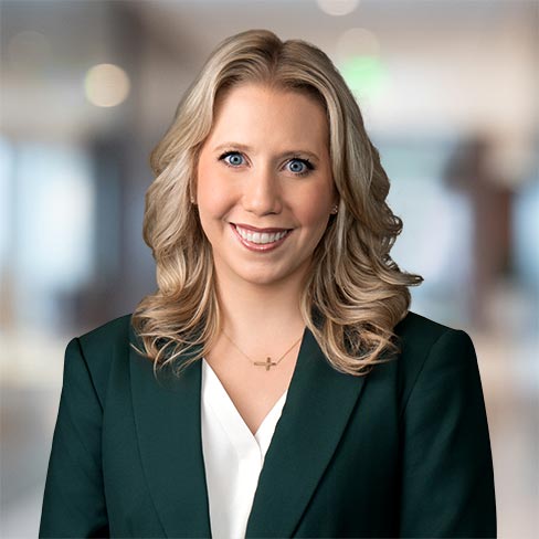 A woman with blonde wavy hair, wearing a dark green blazer and white blouse, smiles at the camera in a blurred office setting.