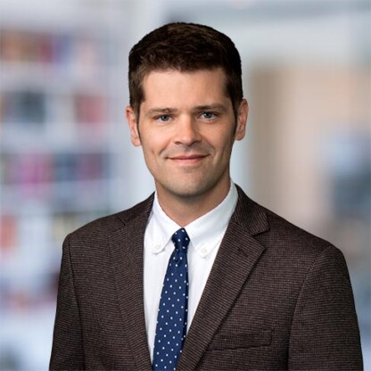 A man in a dark suit and polka dot tie poses for a professional portrait in front of a blurred office background.