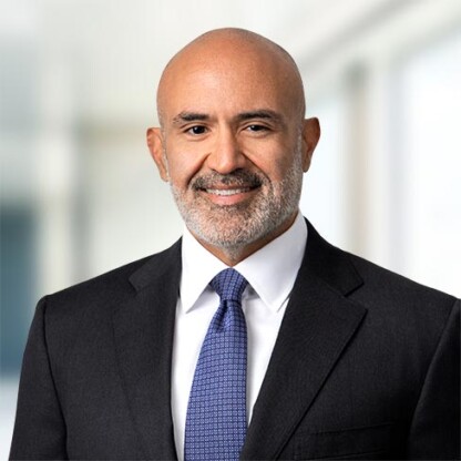 A bald man with a beard, wearing a dark suit, white shirt, and blue tie, stands in front of a blurred office background.