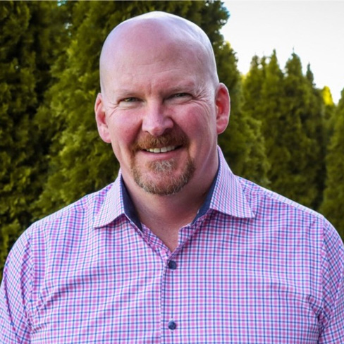 A bald man with a goatee, wearing a pink checkered button-up shirt, smiles outdoors in front of green trees.