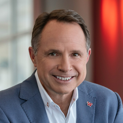 Smiling man in a light blue blazer and white shirt, sitting indoors with blurred background and red lighting.