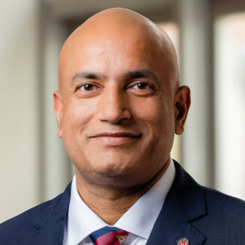 A bald man in a suit and tie smiles slightly while posing for a professional portrait with a blurred indoor background.