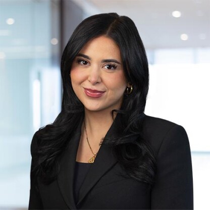 Woman with long black hair wearing a black blazer and gold necklace, standing in a modern office setting with blurred background.