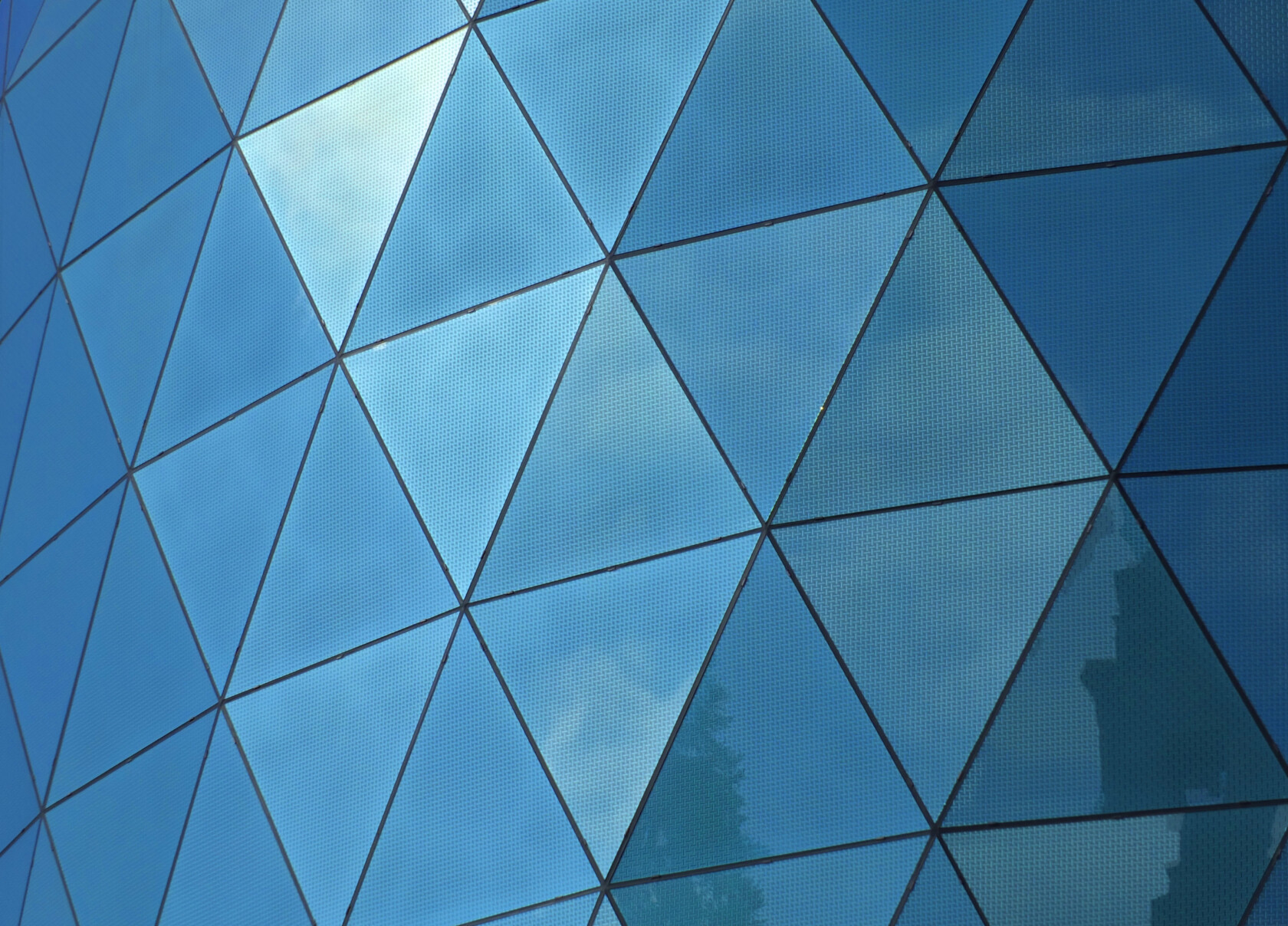Glass building facade with triangular panels reflecting blue sky and clouds.