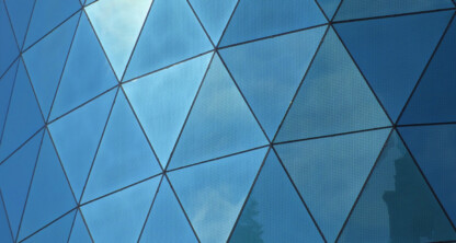 Glass building facade with triangular panels reflecting blue sky and clouds.