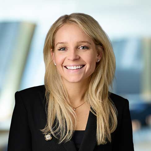 A woman with long blonde hair, wearing a black blazer and smiling, stands in front of a blurred office background.