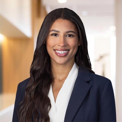 A woman with long dark hair, wearing a navy blazer and white blouse, smiles in a brightly lit office hallway.