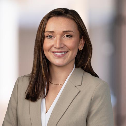 A woman with straight brown hair wearing a beige blazer and white top is smiling at the camera against a blurred office background.
