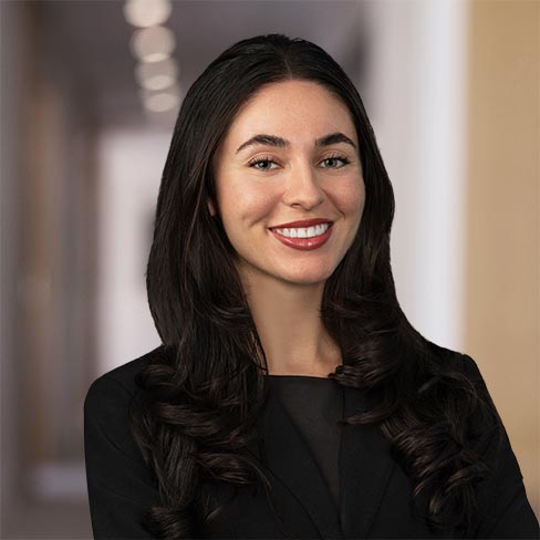 Woman with long dark hair wearing a black blazer and top, smiling, standing in a blurred hallway or office corridor.