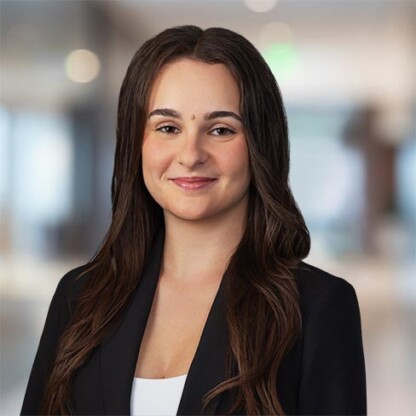 A woman with long brown hair wearing a black blazer and white top, standing in a modern office environment and smiling slightly.