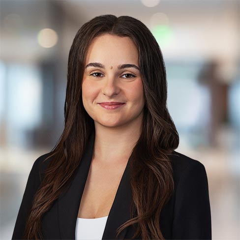 A woman with long brown hair wearing a black blazer and white top, standing in a modern office environment and smiling slightly.