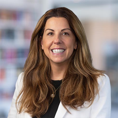 A woman with long brown hair wearing a white blazer and black top smiles at the camera against a blurred indoor background.