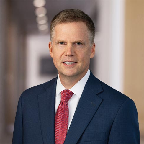A man in a navy blue suit, white shirt, and red tie stands in a blurred hallway, looking at the camera with a neutral expression.