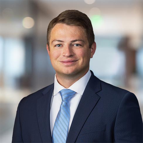 A man in a navy suit, white shirt, and light blue tie poses for a professional headshot in an office setting.