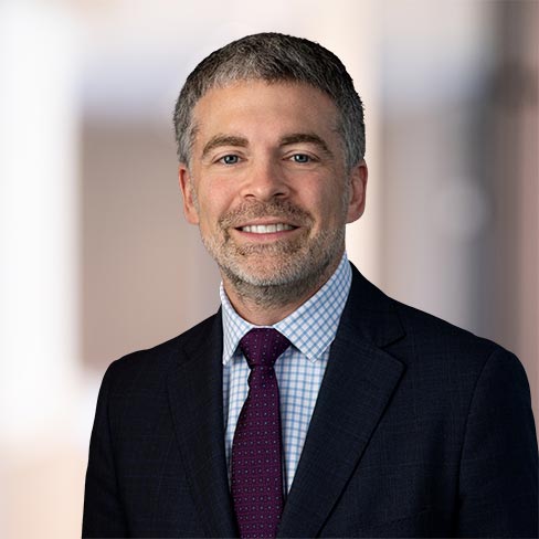 A man in a dark suit, checked shirt, and maroon tie smiles at the camera with a blurred office background.
