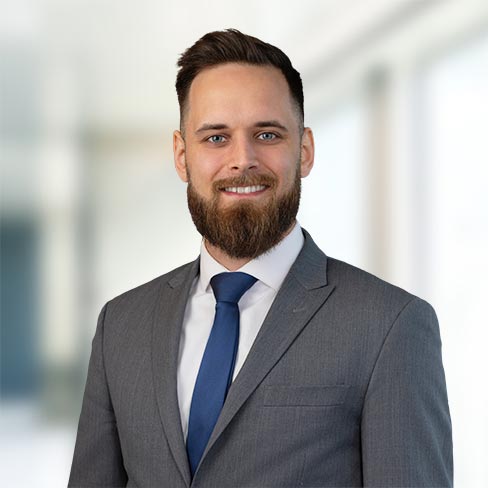 A man with a beard and short hair is wearing a gray suit, white shirt, and blue tie, standing in a bright, blurred office setting.