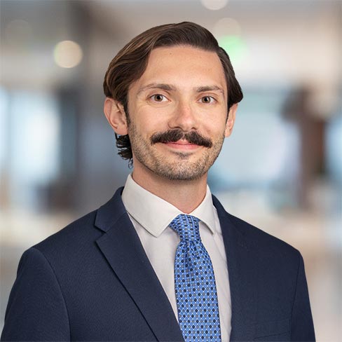 A man with brown hair and a mustache wearing a navy suit, white shirt, and blue patterned tie stands in an office setting.