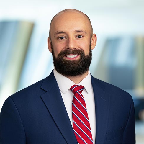 A man with a bald head and full beard wearing a blue suit, white shirt, and red striped tie stands in front of a blurred office background.