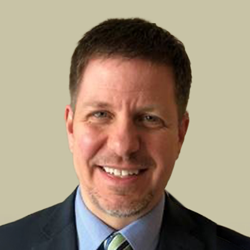 A man with short brown hair and a trimmed beard, wearing a suit, light blue shirt, and striped tie, smiling at the camera against a plain beige background.