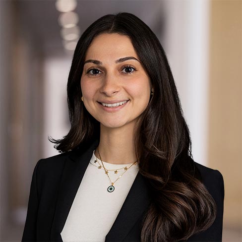 A woman with long dark hair, wearing a black blazer, white top, and layered necklaces, smiles at the camera in a well-lit hallway.