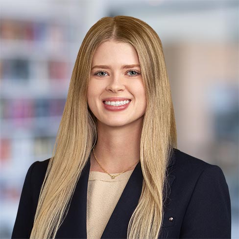 A woman with long blonde hair, wearing a dark blazer over a beige top, smiles at the camera in an office setting with blurred shelves in the background.