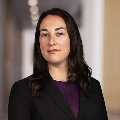 Woman with long dark hair wearing a black blazer over a purple top, standing in a professional indoor setting with blurred background.