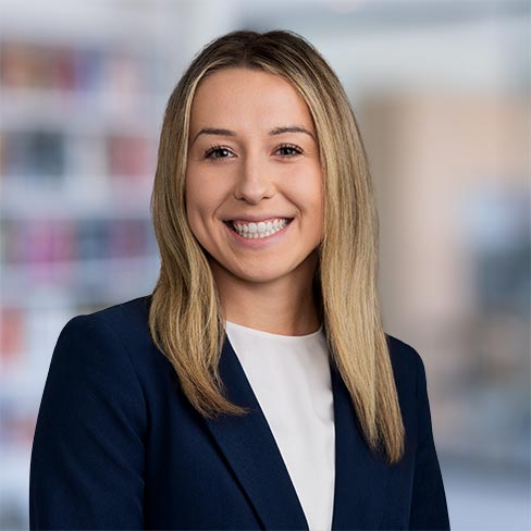 A woman with straight blonde hair, wearing a navy blazer and white top, smiles at the camera against a blurred office background.