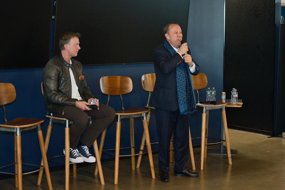 Two men are on a stage; one is seated holding a drink, while the other stands speaking into a microphone. Four wooden chairs and water bottles are visible.