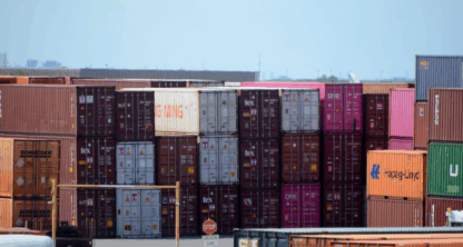 Stacked shipping containers in various colors are organized at a shipping yard, with a stop sign and traffic signs visible in the foreground.
