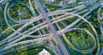 Aerial view of a complex highway interchange with multiple lanes, overpasses, and looping ramps surrounded by greenery and nearby buildings.